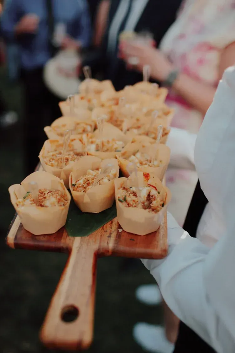 Tray of bite-sized appetizers at a buffet, with cups of creamy topping and crispy garnish being carried by a server
