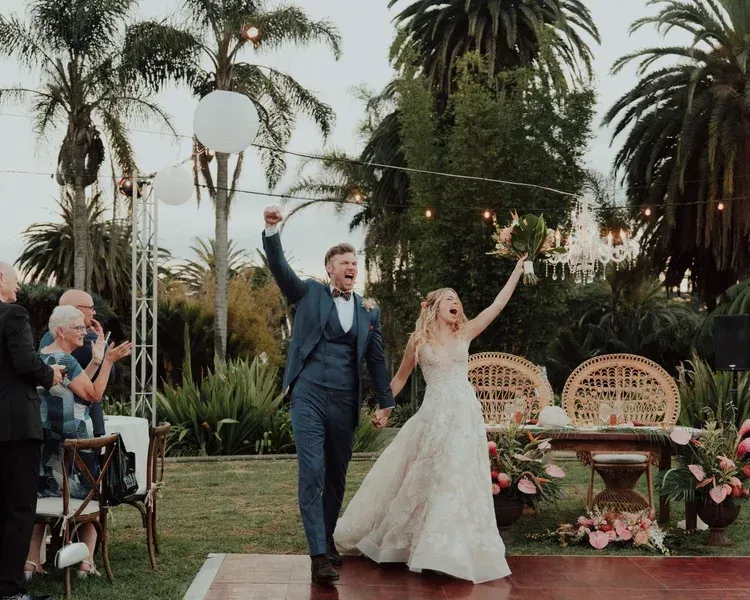 Bride and groom celebrating outdoors in a garden, raising their arms beside floral tables and palm trees.