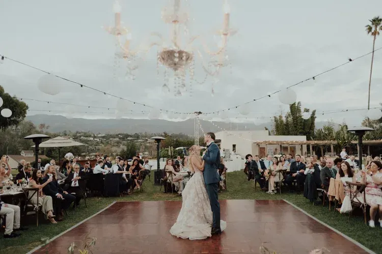 Wedding couple dancing on an outdoor wooden floor under a chandelier at a reception with seated guests