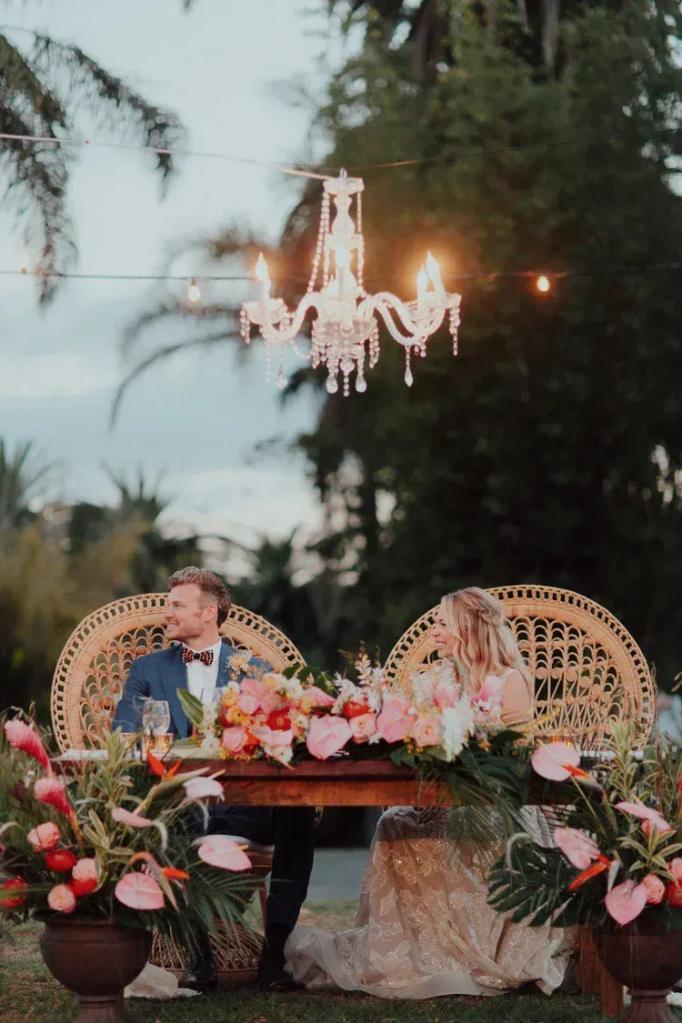 Outdoor wedding reception with chandelier lighting, floral decor, and a couple seated at a table by the water.