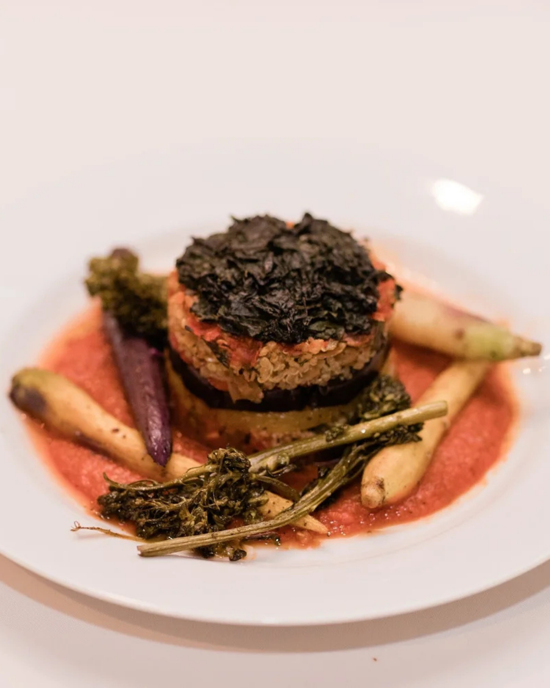 Seared meat medallion with dark herb crust, roasted vegetables, and red sauce on a white plate