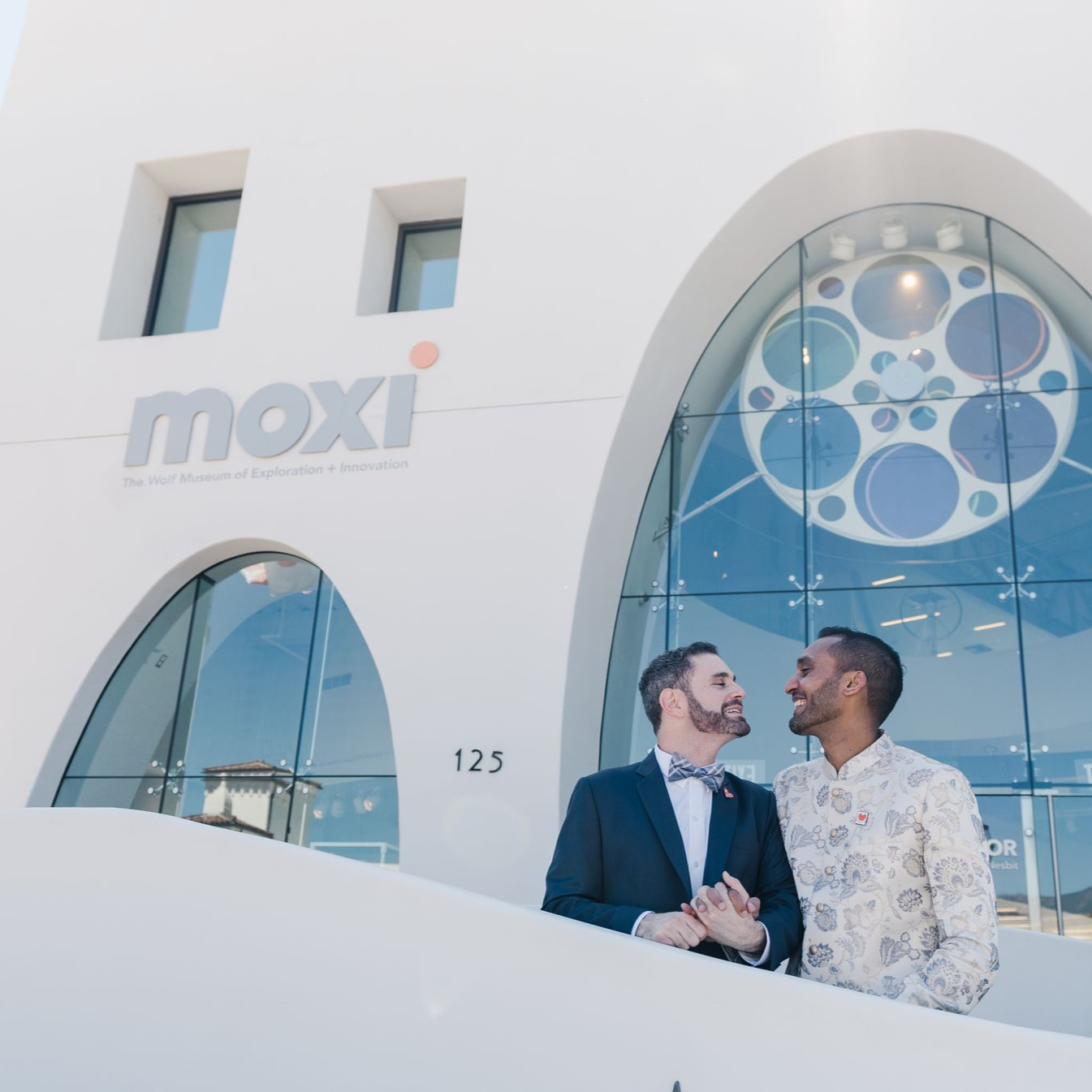 Two people smiling and holding hands outside a white building with large circular windows and a “moxi” sign
