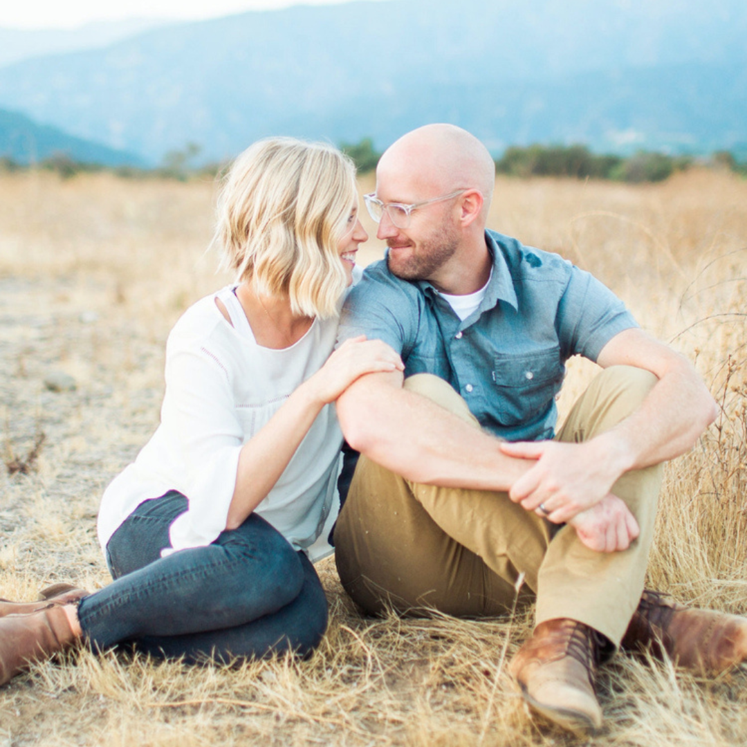 Two people sitting in dry grass, smiling at each other with mountains in the background