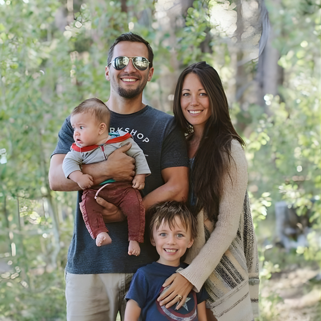 Family of four smiling outdoors, with parents holding a baby and young child in front.