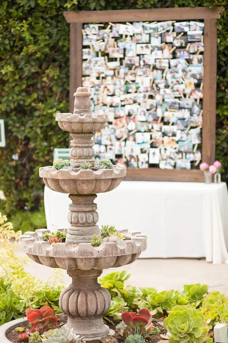 Three-tier stone fountain before a framed photo display and white table outdoors