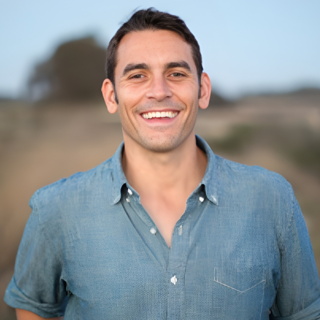 Smiling man in a blue shirt outdoors with a blurred landscape background