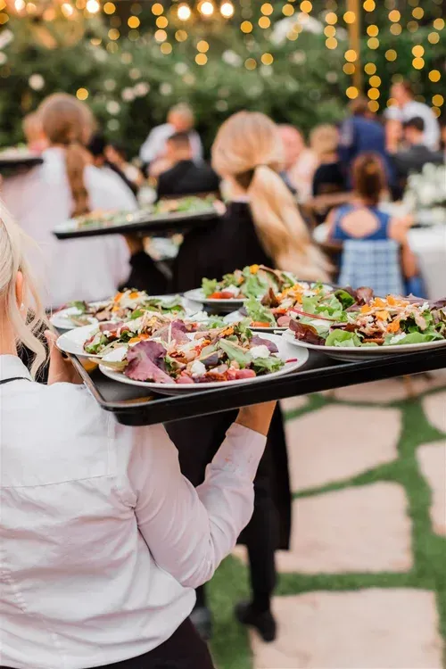 Server carrying trays of colorful salads at an outdoor evening event with string lights