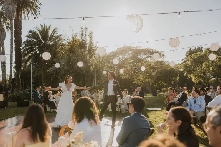 Wedding couple dancing outdoors at sunset with guests seated around them and hanging lanterns overhead