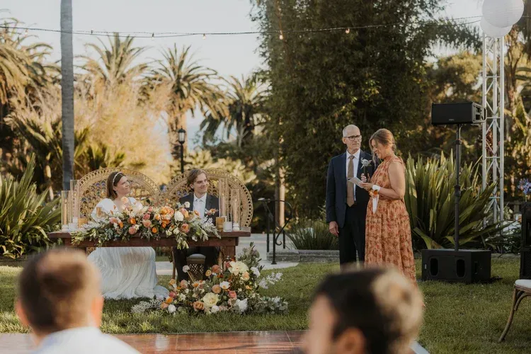 Outdoor wedding ceremony with couple at the altar and officiant under palm trees, floral arrangements nearby