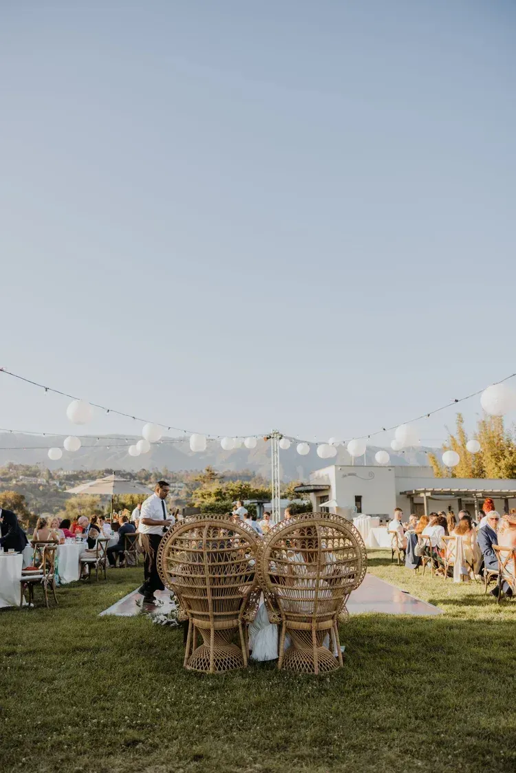 Outdoor lawn event with wicker chairs, string lights, and guests in the background.