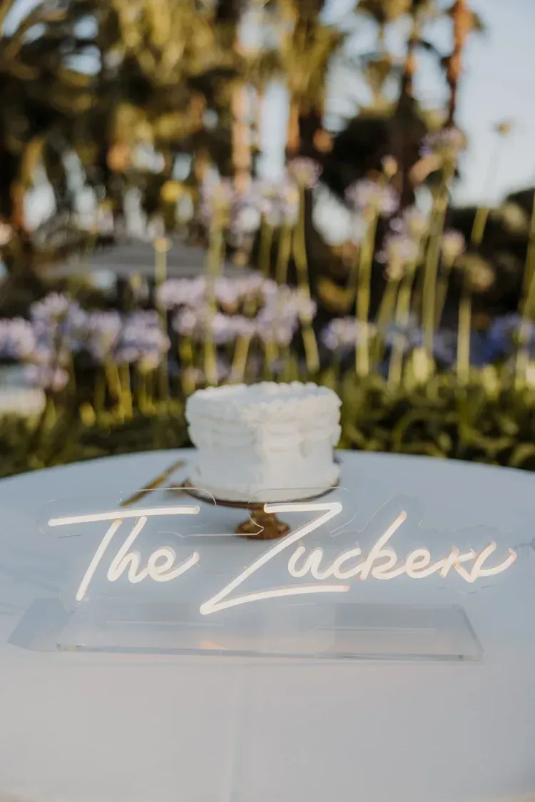 White cake on a clear stand with a neon-style “The Zucker” sign outdoors, blurred palm trees behind