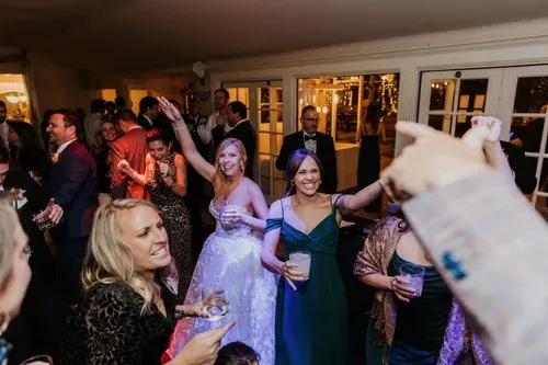 Guests dancing and smiling at a lively indoor party, with a woman holding a drink in the foreground.