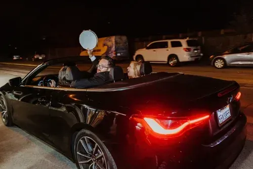 Black convertible at night with two passengers, red taillights glowing on a city street.