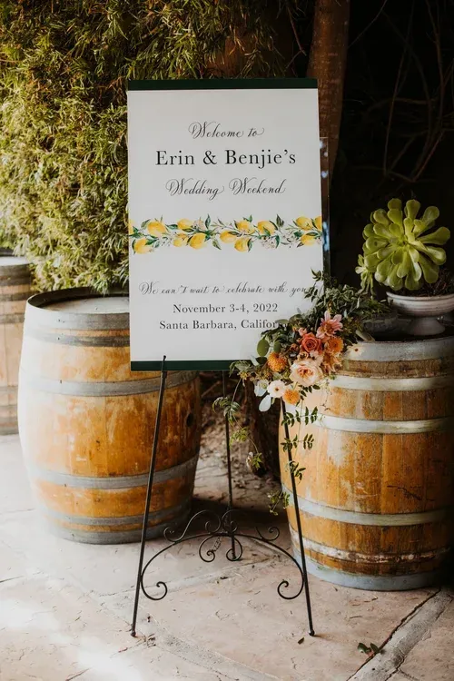 Wedding welcome sign on easel, flanked by wooden barrels and floral arrangements outdoors.