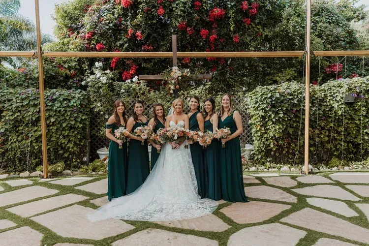Bridal party posing in a garden, bride in a white gown with bridesmaids in dark green dresses.
