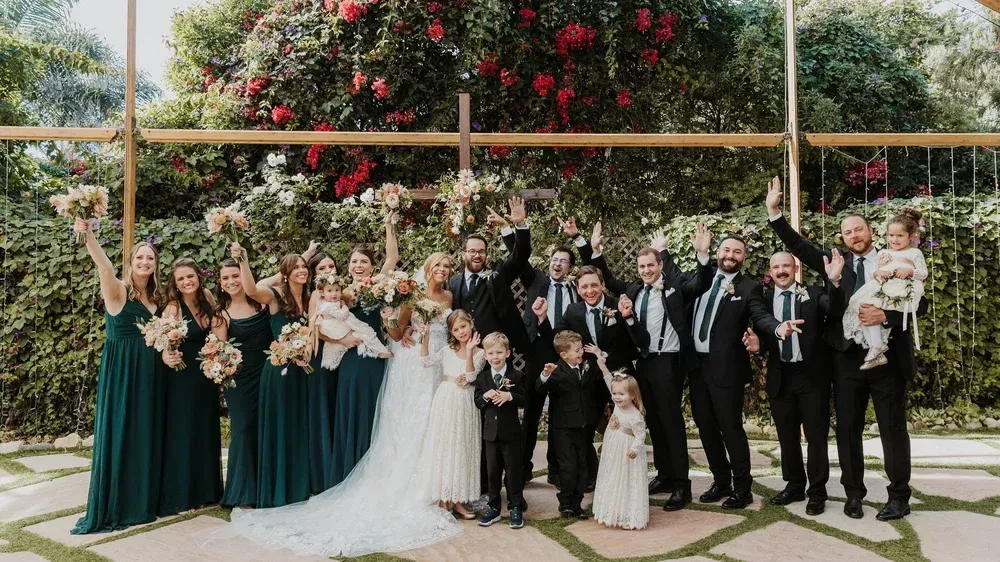 Wedding party posing outdoors under a floral arch, with the bride in white and attendants in dark green and black attire