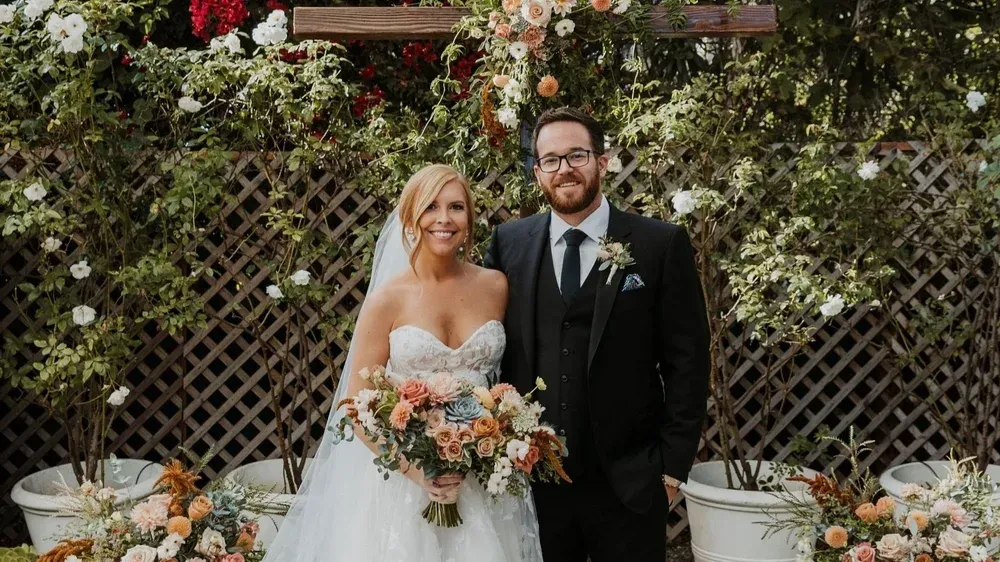 Bride and groom stand smiling in a garden wedding setting, holding a bouquet