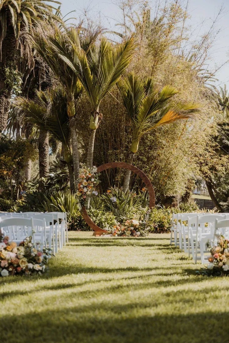Outdoor wedding aisle with white chairs, floral arrangements, and palm trees in a sunny garden