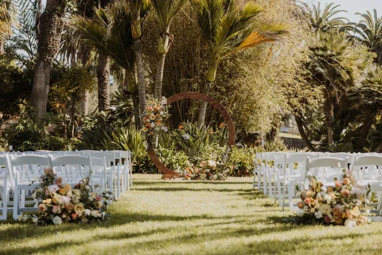 Outdoor wedding setup with white chairs, floral arrangements, and palm trees in a garden lawn