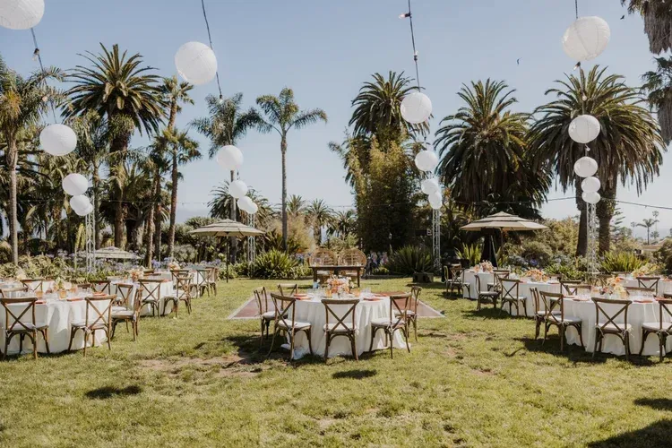 Outdoor garden reception with round tables, white chairs, palm trees, and hanging lanterns