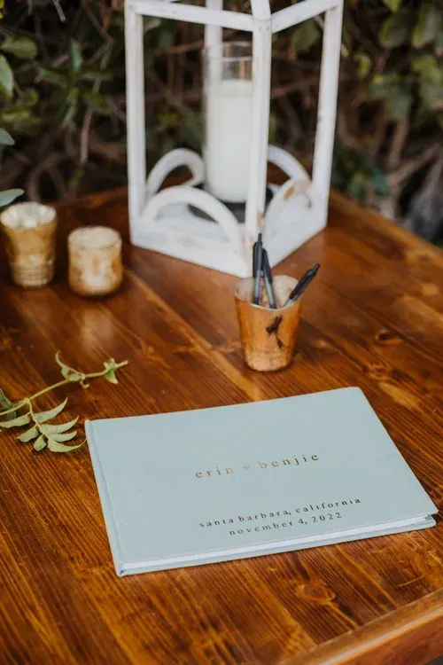 Blue wedding guestbook on a wooden table beside a small lantern and candle holders.