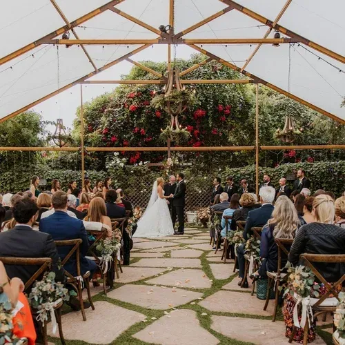 Outdoor wedding ceremony under a greenhouse-like pavilion, bride and groom at the altar, guests seated on both sides