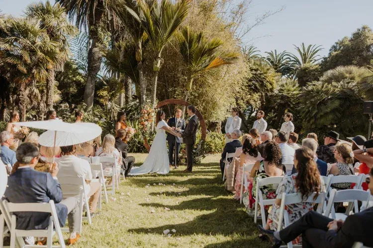 Outdoor wedding ceremony in a garden, with bride and groom standing beneath an arch among seated guests.