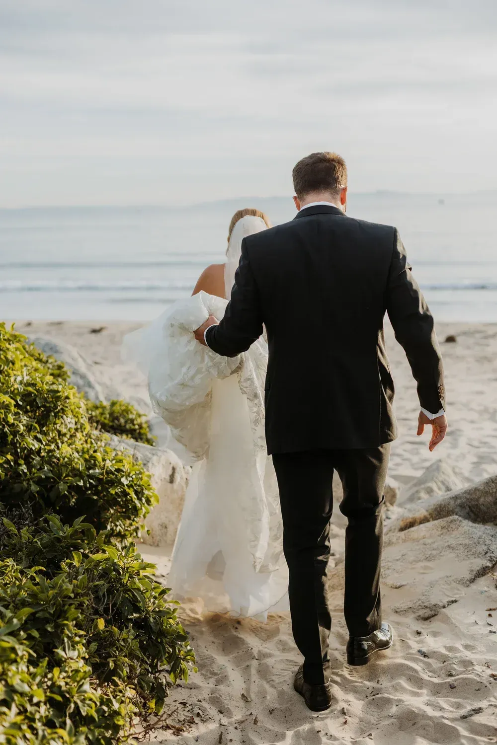 Couple in wedding attire walking toward the beach on a sandy path at sunset