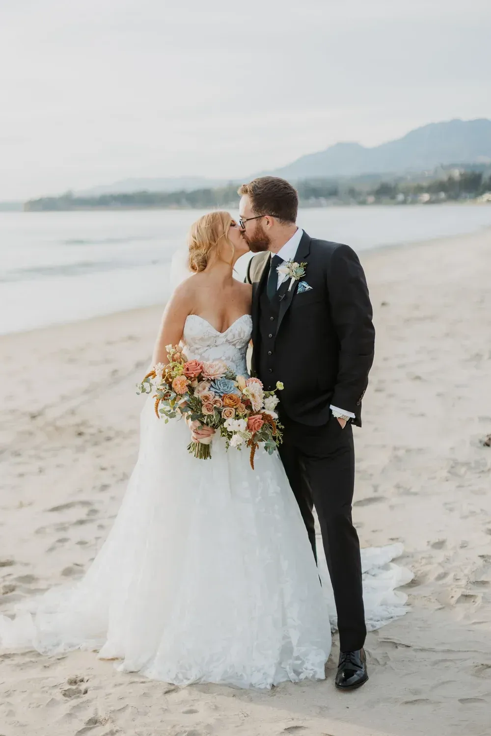 Bride and groom kissing on a beach, bride holding a colorful bouquet, with mountains in the background
