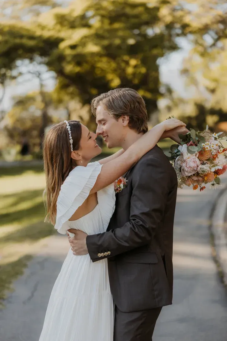 Couple embracing and kissing outdoors, the woman in a white dress holding a bouquet.
