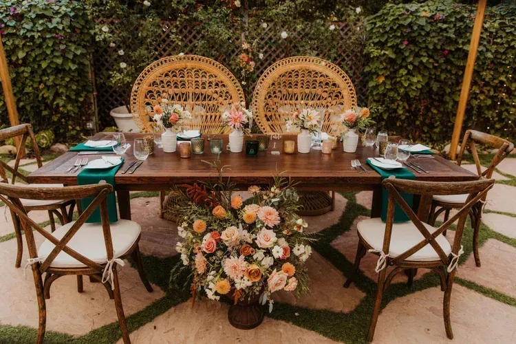Outdoor table set for a garden dinner, with wicker chairs, flowers, candles, and greenery backdrop