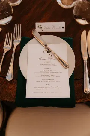 Elegant place setting with white menu, gold utensils, and a dark green napkin on a wooden table
