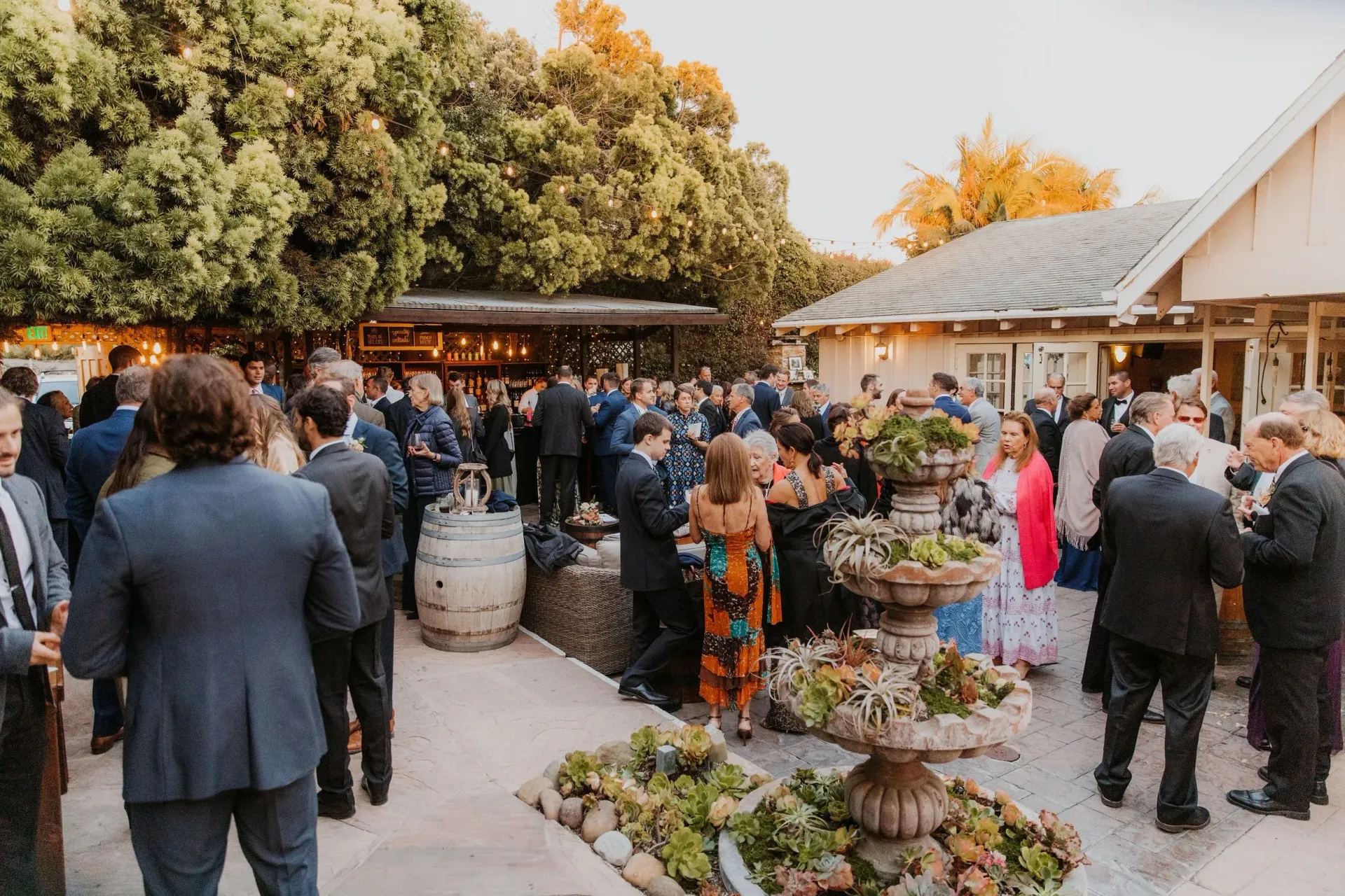 Crowd mingling in an outdoor market garden with potted plants and trees under warm evening light
