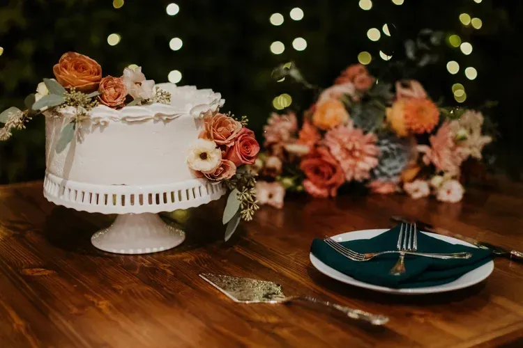 Elegant white wedding cake with peach roses on a wooden table beside a teal plate and floral centerpiece