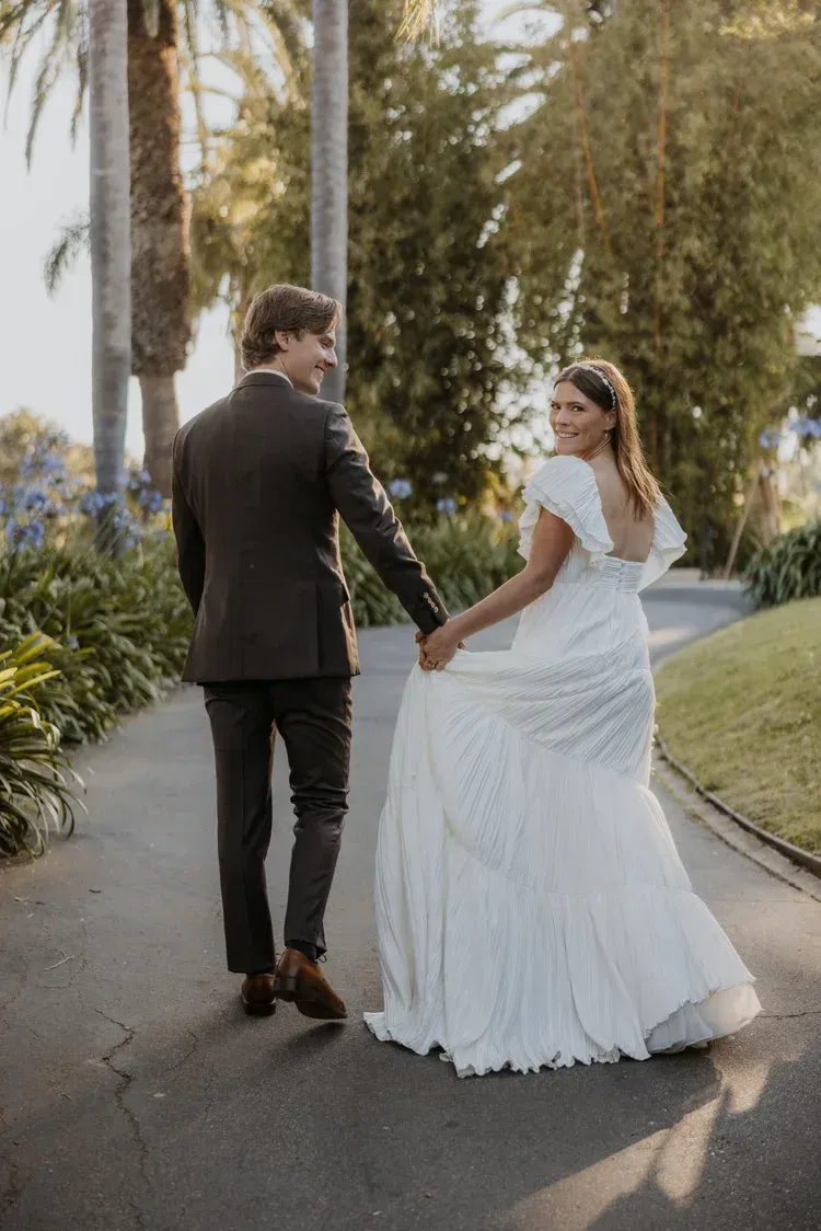 Newlyweds holding hands and walking down a palm-lined path outdoors at sunset