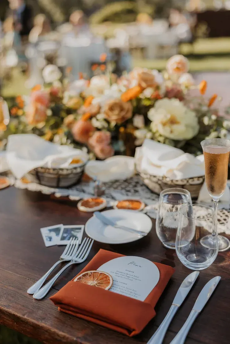 Elegant outdoor dining table with orange napkins, flowers, wine glasses, and place settings