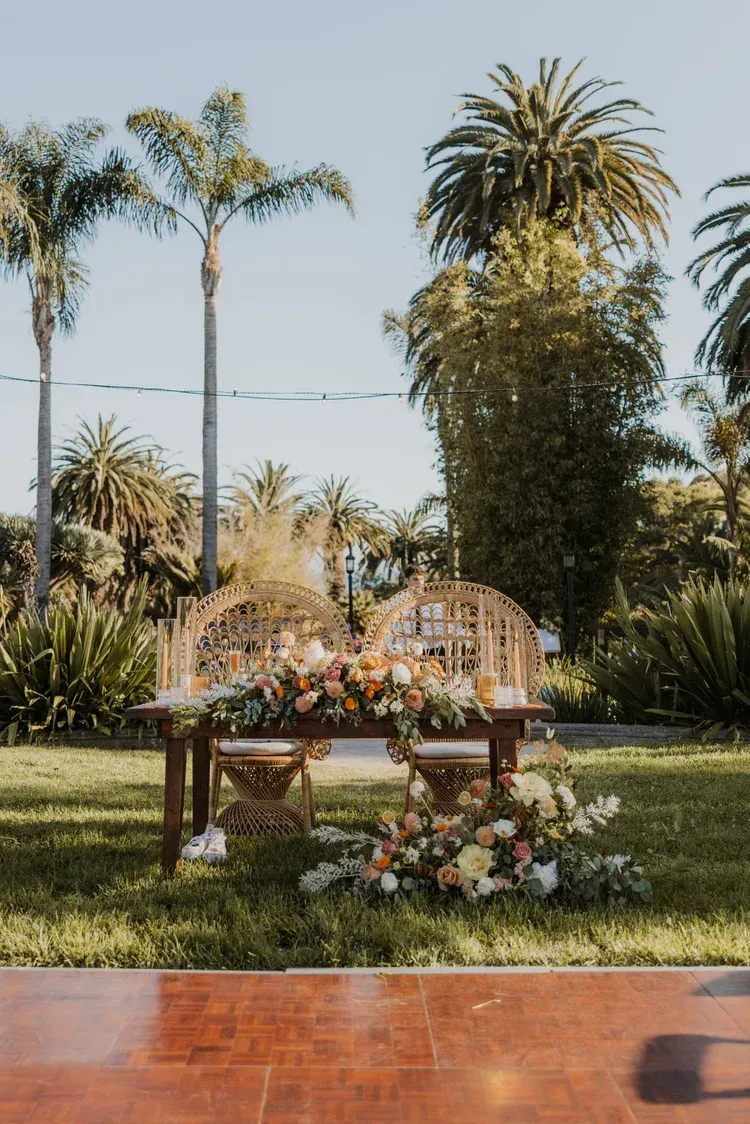 Decorated outdoor table set on a lawn with flowers, framed by palm trees in a sunny garden.