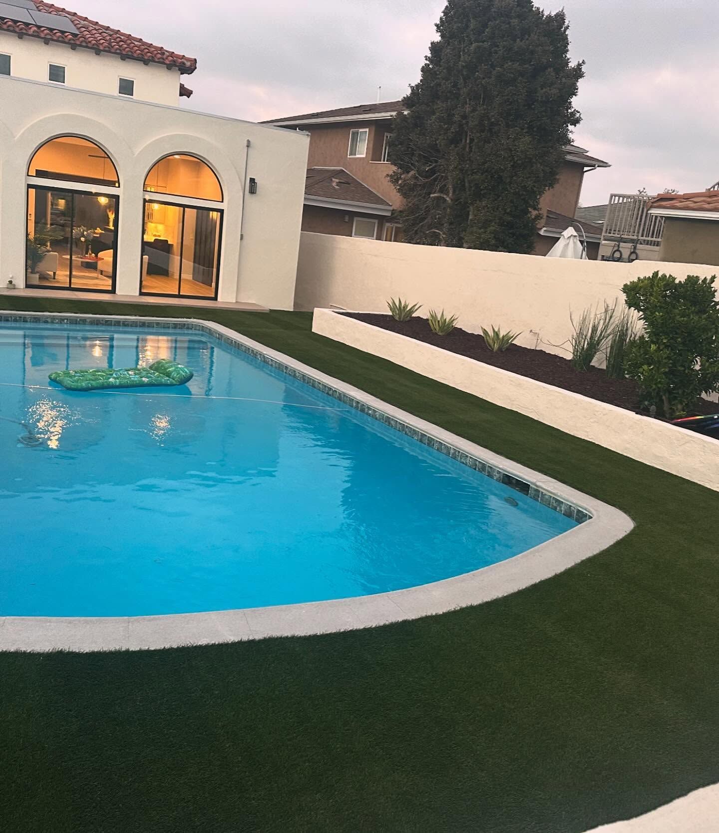 Backyard swimming pool beside a house with arched windows at dusk, surrounded by grass and a white wall
