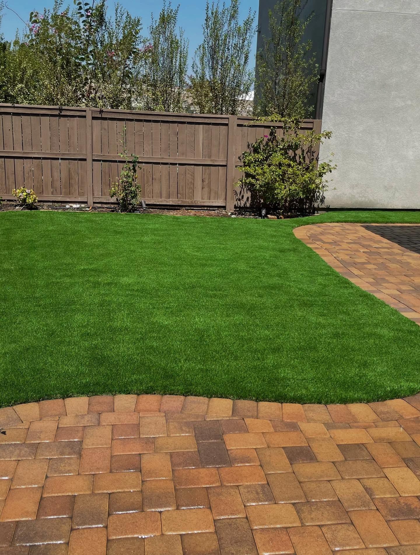 Backyard with green lawn, brick patio, wooden fence, and a small shrub by the wall