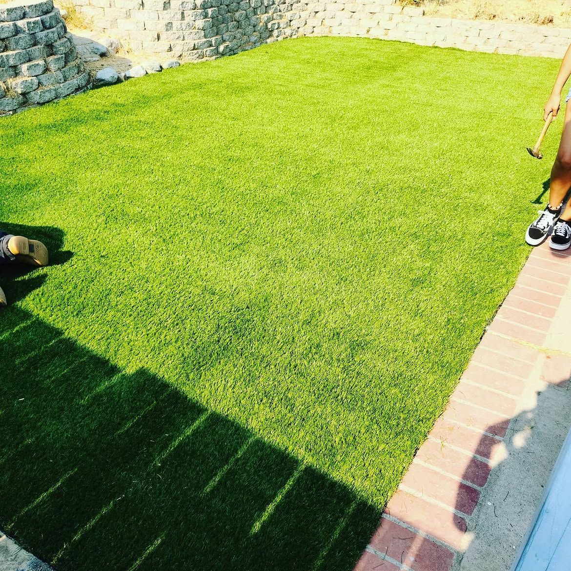 Sunny backyard with green lawn, stone wall, brick path, and a person partly visible at the right edge