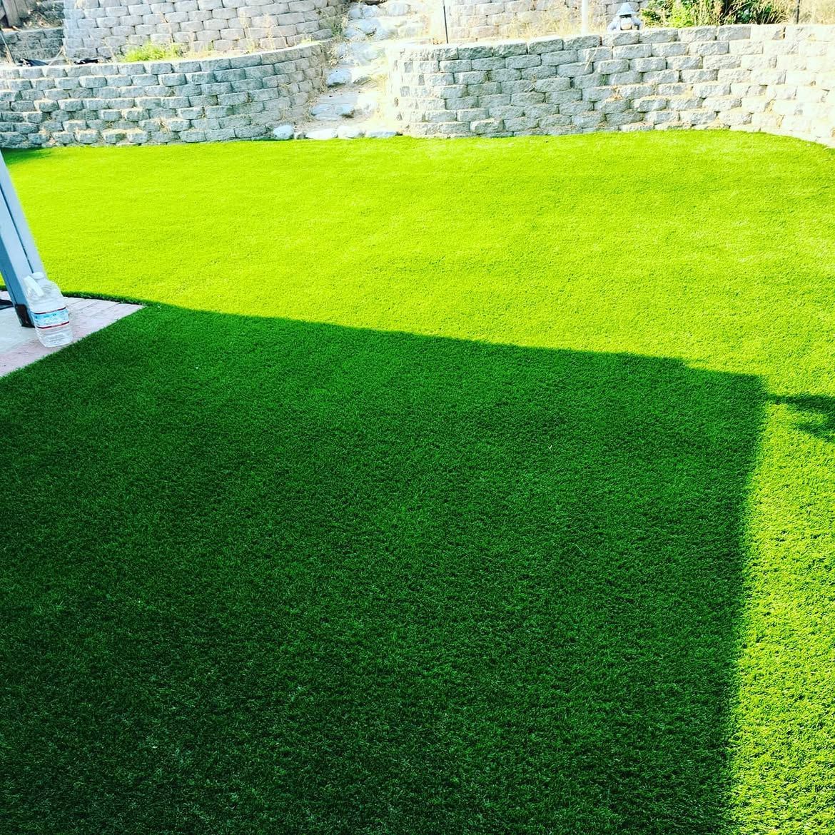 Bright green artificial turf with a large shadow beside a stone wall and blue post