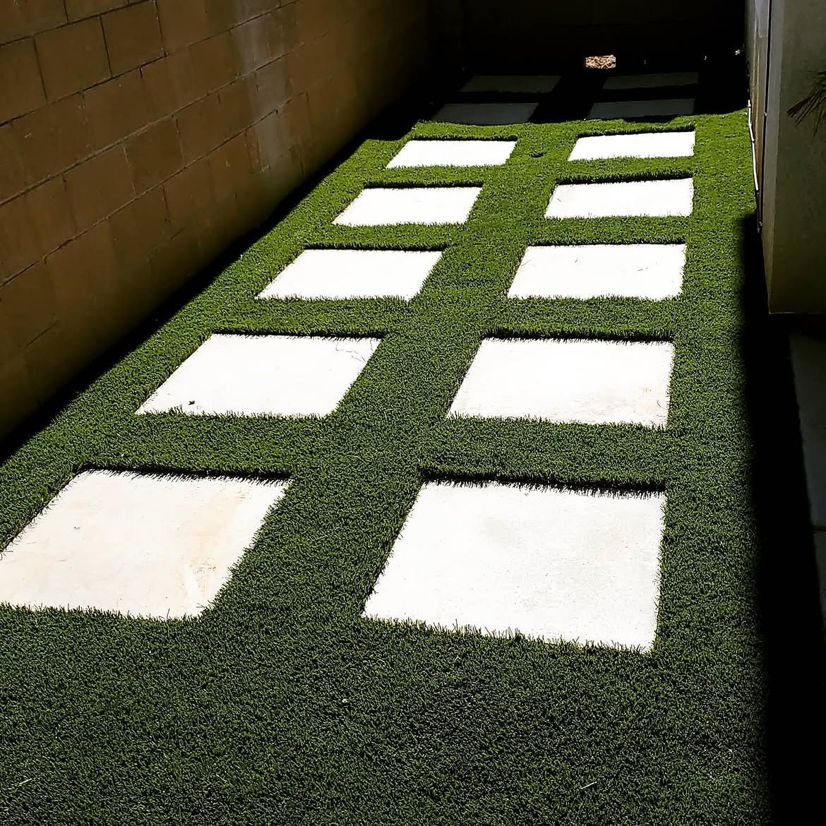 Artificial turf walkway with white square stepping stones in a narrow, sunlit corridor