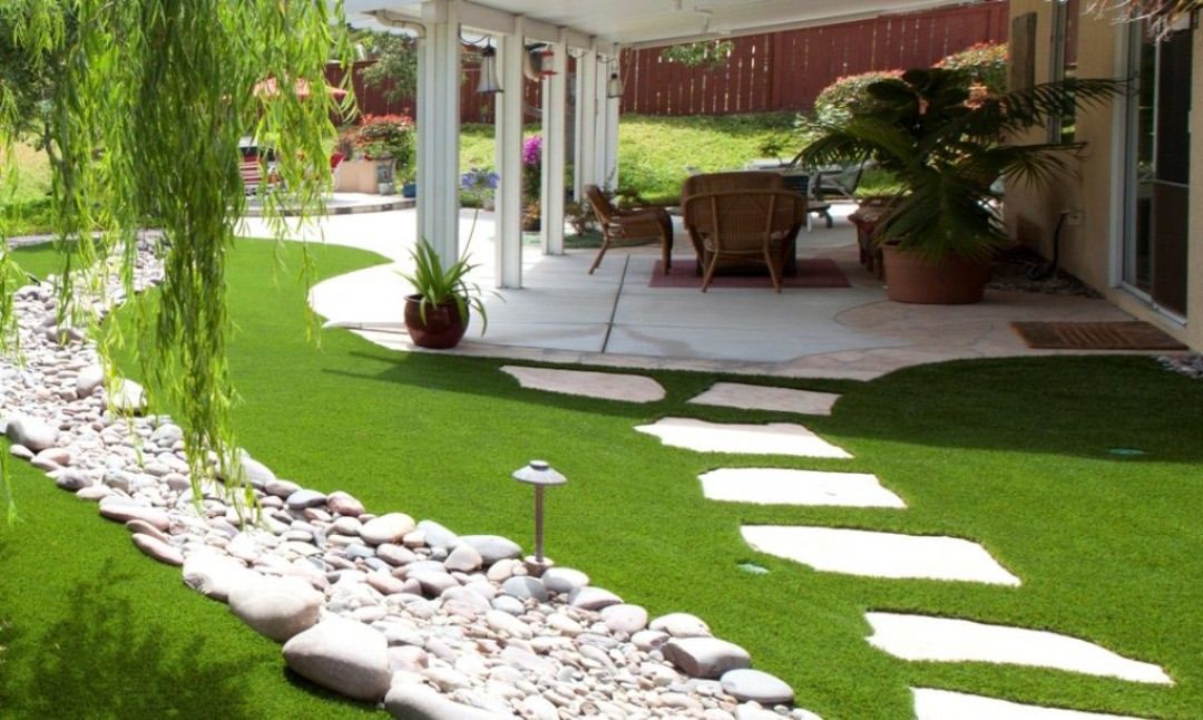 Sunny backyard patio with white pavers, green lawn, stone border, hanging plants, and outdoor seating.