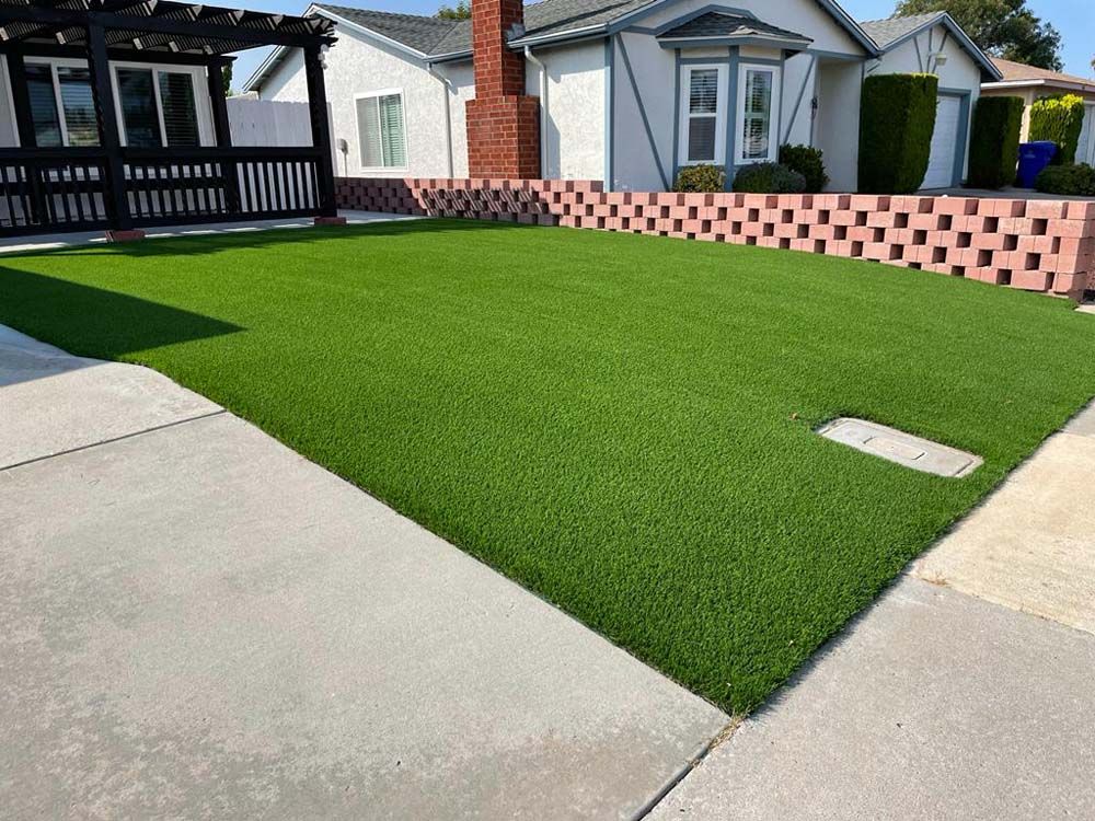 Backyard with bright green artificial turf, concrete patio, and a pink brick retaining wall with houses behind it