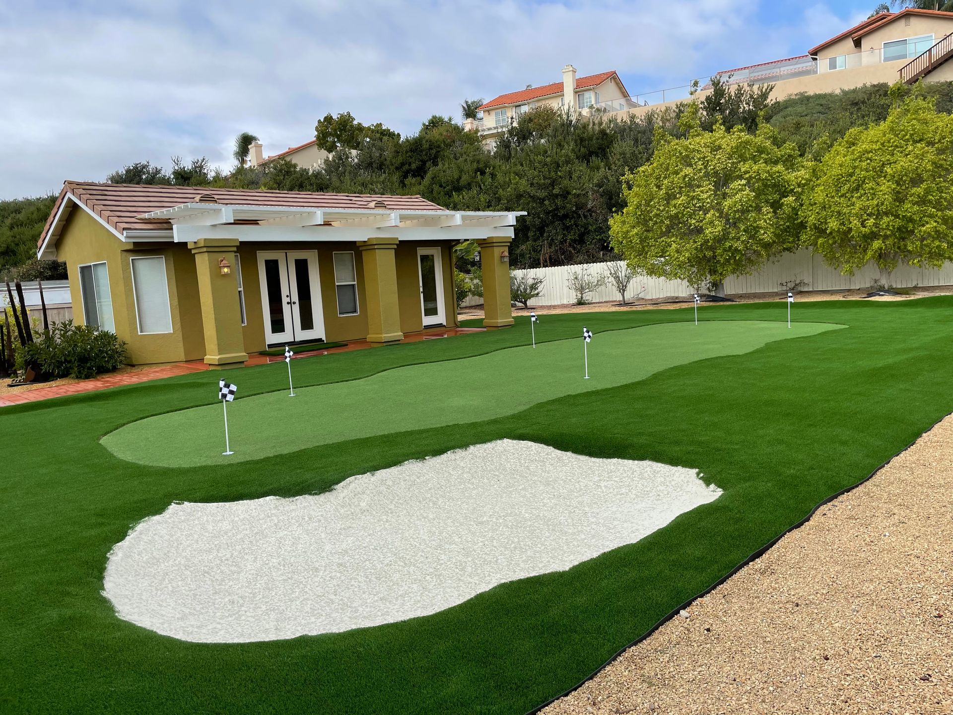 Backyard putting green with sand bunker, small house, and hillside homes in the background