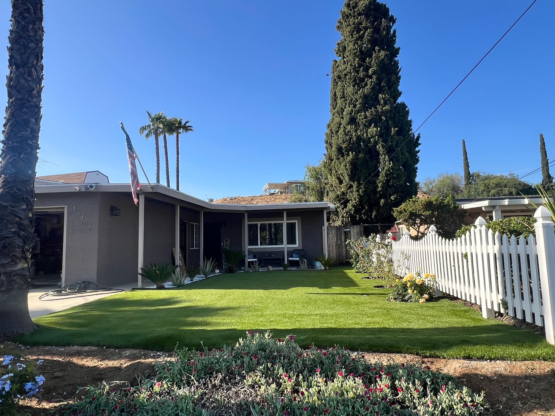 Single-story house with a green lawn, white picket fence, and tall cypress trees under a clear blue sky