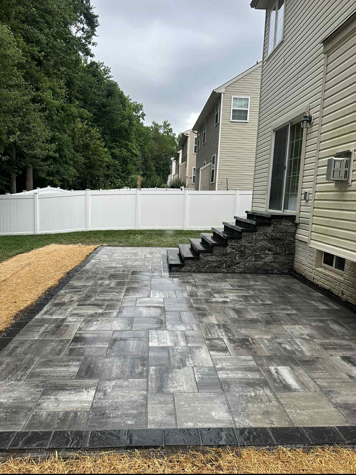 A paved patio with dark gray pavers, steps leading to a back door, and a white fence in a backyard setting. Perry Hall, MD - Oakwood Landscaping LLC