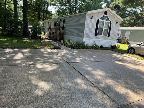 White mobile home with damaged siding beside a concrete driveway in a wooded area