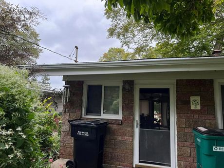 Small brick shed with white-trimmed windows, black door, and trash bins under trees.