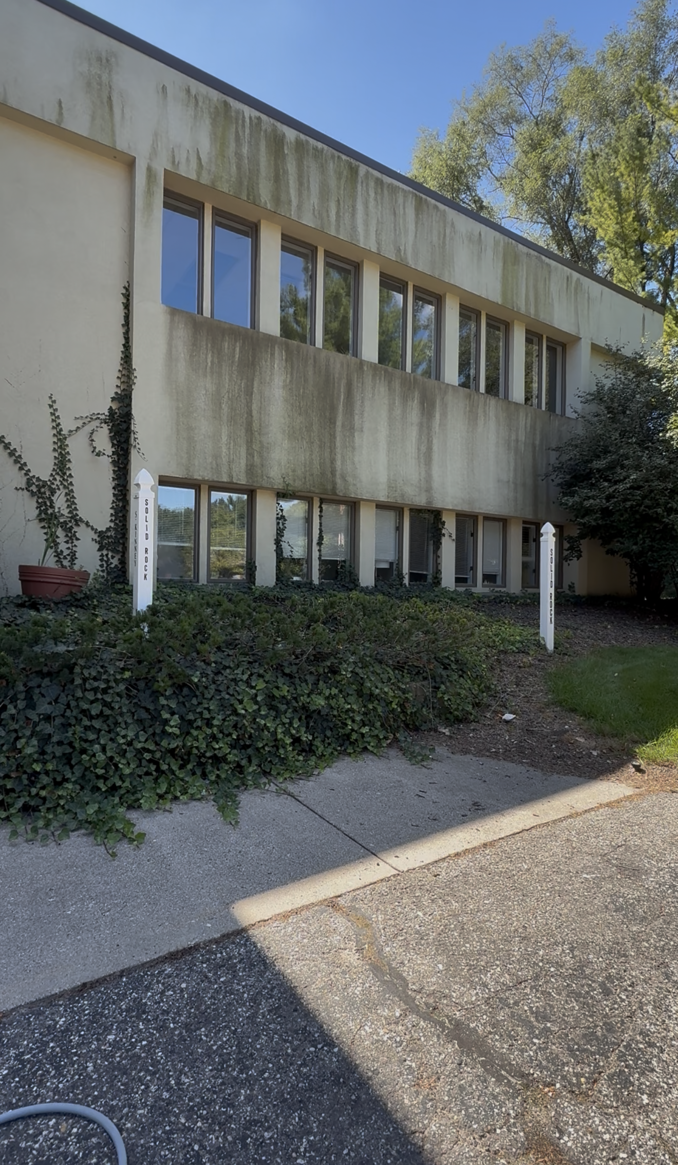 Two-story beige office building with ivy and shrubs, viewed from a paved path under a blue sky